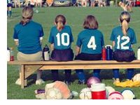 Rückansicht: Mädchen in blauen Fußballtrikots und Trainerin sitzen nebeneinander auf einer Bank am Rand eine Fußballfeldes.
