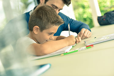 Vergrößerungsansichten für Bild: Nahaufnahme: Ein Junge macht seine Hausaufgaben und ein Erwachsener sitzt helfend daneben.