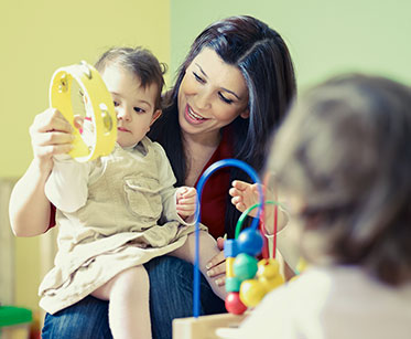 Vergrößerungsansichten für Bild: Nahaufnahme: Mitarbeiterin einer Kinderkrippe beim Spielen mit einem kleinen Kind, das mit einer Rassel auf ihrem Schoß sitzt. 